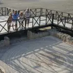 People stand on a wooden platform in Western Cyprus, overlooking an ancient mosaic nestled among stone ruins. This site, rich with living traditions, offers a glimpse into the vibrant history that shaped this land.