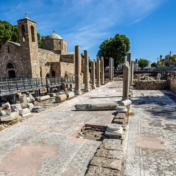 Ancient ruins with mosaic floor and scattered columns; a stone church stands in the background under a clear blue sky.