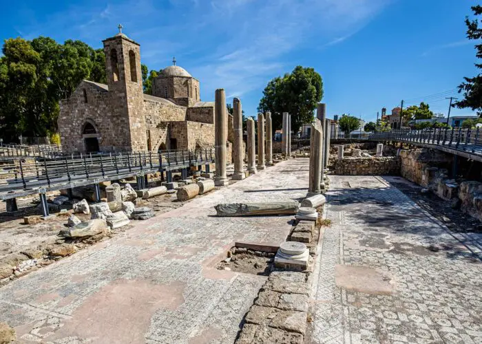 Ancient ruins with mosaic floor and scattered columns; a stone church stands in the background under a clear blue sky.