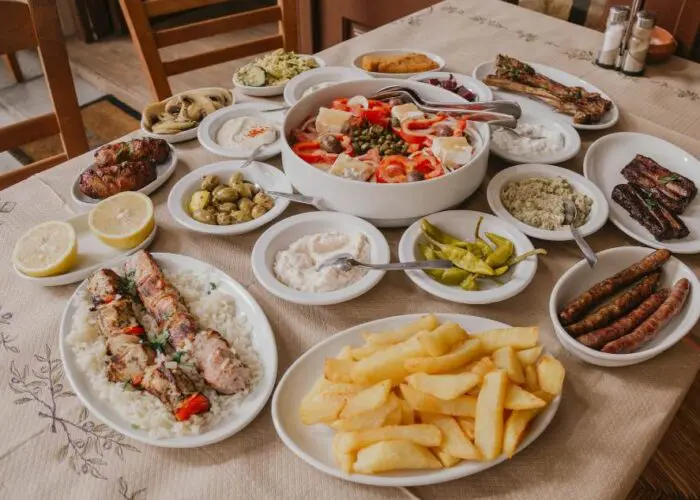A table filled with various dishes including grilled meats, French fries, vegetable salads, dips, olives, bread, and lemon slices, displayed on white plates and bowls.