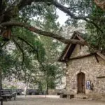 A small stone building with wooden doors and windows stands under large, old trees with a bench and framed pictures nearby, against a backdrop of hills and greenery.