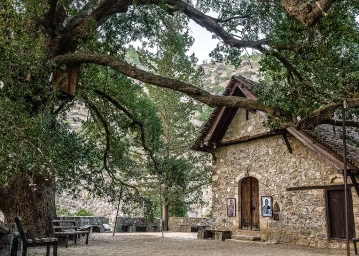 A small stone building with wooden doors and windows stands under large, old trees with a bench and framed pictures nearby, against a backdrop of hills and greenery.