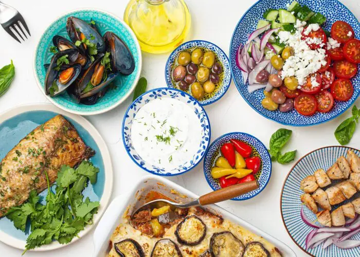 A variety of Greek food dishes including grilled fish, mussels, a Greek salad, olives, sauce, grilled meat, and eggplant are arranged on a table.