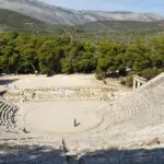 Ancient amphitheater with tiered stone seating and a central stage, surrounded by forested hills and mountains. Sparse visitors are scattered around the seating area and stage.