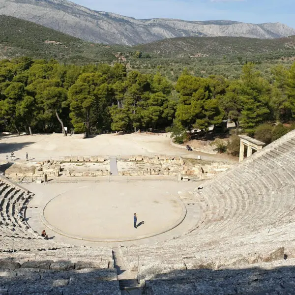 Ancient amphitheater with tiered stone seating and a central stage, surrounded by forested hills and mountains. Sparse visitors are scattered around the seating area and stage.