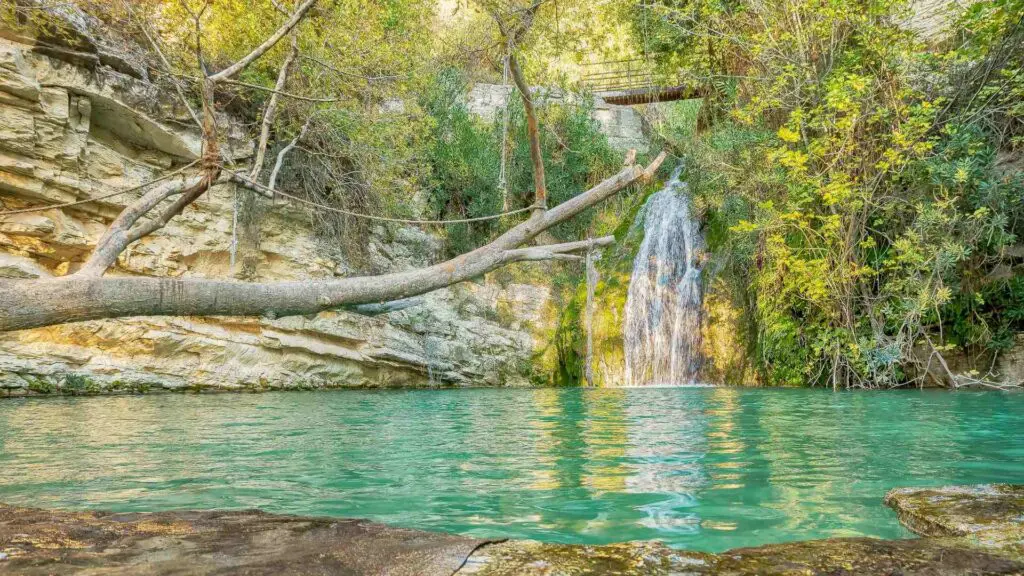 A waterfall flows into a turquoise pool surrounded by rocky cliffs and lush greenery, with a log extending over the water. This natural wonder resembles a scene from mythology, nestled within the serene landscapes of Akamas National Park.