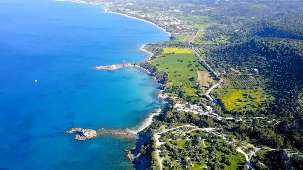 Aerial view of the Akamas National Park reveals a coastal landscape with clear blue water, rocky shorelines, and lush green vegetation. A winding road stretches along the coast, making it a perfect scene for a travel guide showcasing natural wonders.