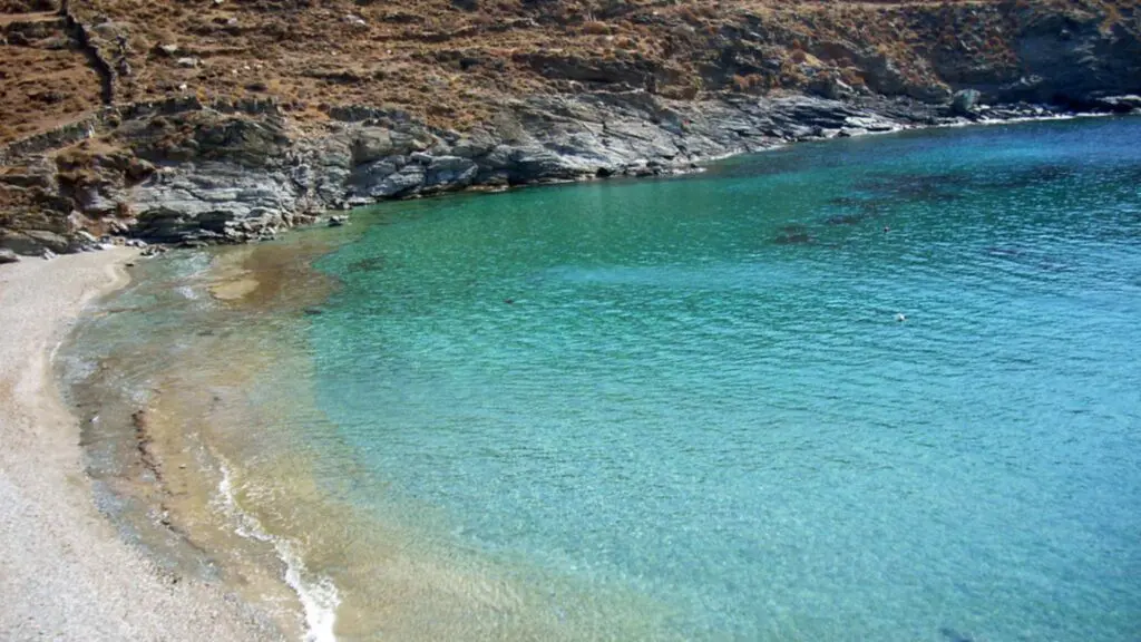 A clear turquoise bay curves against a rocky hillside with a narrow sandy beach in the foreground, welcoming visitors to the serene beauty of Kythnos Island.