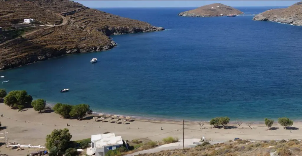 Aerial view of a beach with umbrellas, trees, and a few small buildings. The coastline features rocky hills, perfect for a travel guide to Kythnos Island. A boat is visible in the water near a distant island.