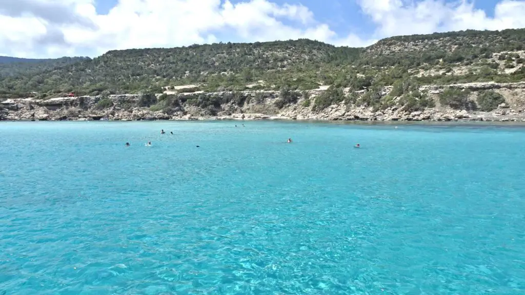 A clear turquoise sea with a few distant swimmers, backed by the rugged cliffs and green hills of Akamas National Park under a partly cloudy sky. This natural wonder is a must-see in any travel guide exploring the region's breathtaking beauty.