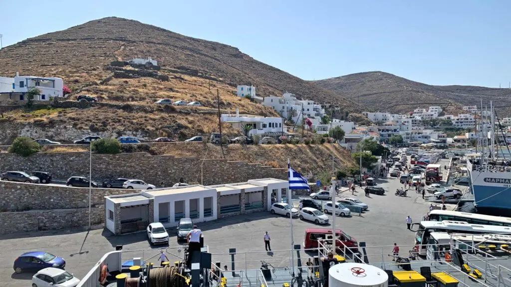 A bustling harbor on Kythnos Island, where parked cars and boats create a lively scene. In the background, hills adorned with white buildings paint a picturesque view, while the Greek flag flutters proudly—a perfect snapshot for any travel guide exploring Kythnos.