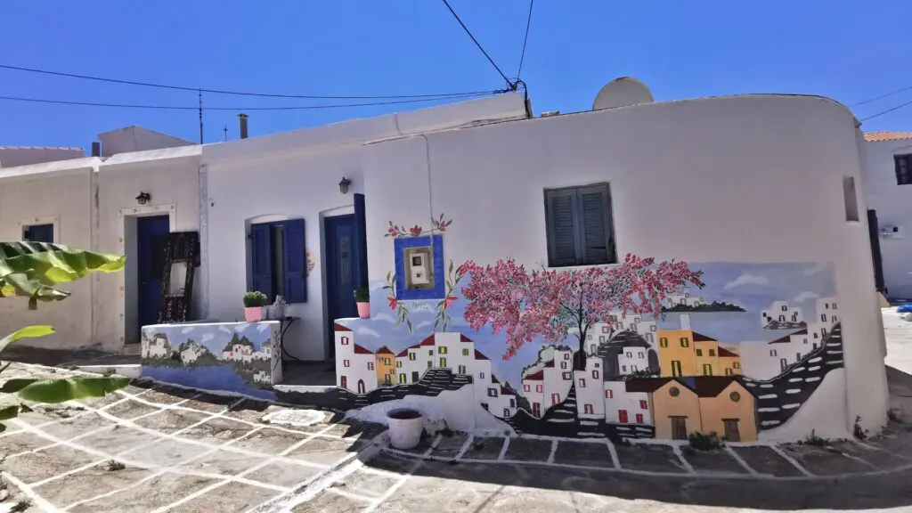 A white building on Kythnos Island features a colorful mural depicting houses and trees. The structure boasts blue doors and windows, perfectly complementing the clear blue sky above, making it a picturesque highlight for any travel guide.