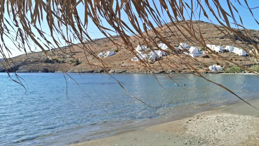 View from a beach on Kythnos Island with clear blue water and sandy shore, a straw umbrella in the foreground, and white buildings perched on a hill in the background—a perfect scene for your travel guide.