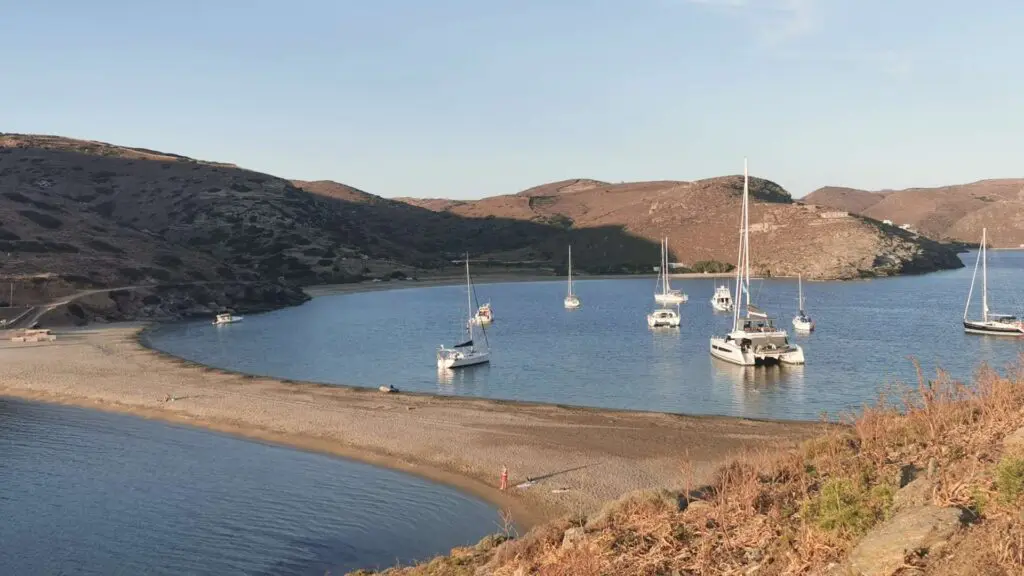 Boats anchored in a calm bay on Kythnos Island are surrounded by hills, with a sandy strip and sparse vegetation nearby under a clear sky.