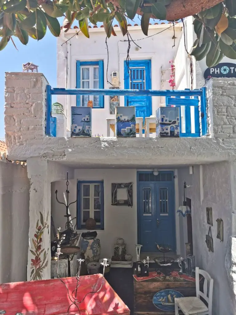 A white two-story building with blue doors and balcony features, adorned with small decorative items, typical of Kythnos charm. A tree's branches partially frame the scene, and a red table is visible in the foreground, perfect for a travel guide’s enticing snapshot of island life.