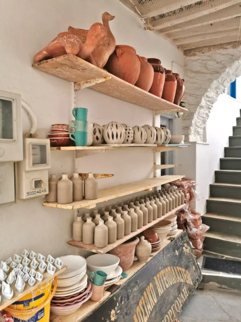 Shelves displaying pottery and ceramics of various shapes and sizes evoke memories of Kythnos Island in a rustic setting with stairs nearby.