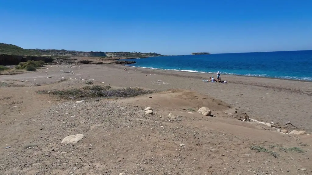 A rocky beach with a few people sitting on the sand, embraced by the clear blue sea and sky at Akamas National Park. Sparse vegetation is visible along the shoreline, adding to this natural wonder's serene beauty.