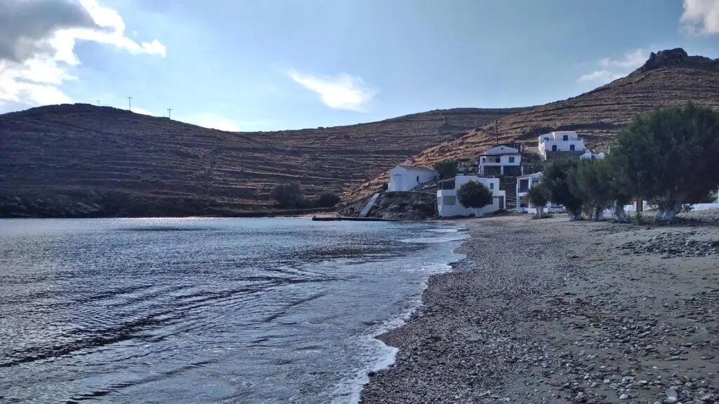 Discover the serene beauty of Kythnos, where a quiet beach with gentle waves is bordered by a hillside and a cluster of white buildings and trees in the background—a perfect spot for any travel guide exploring this enchanting island.