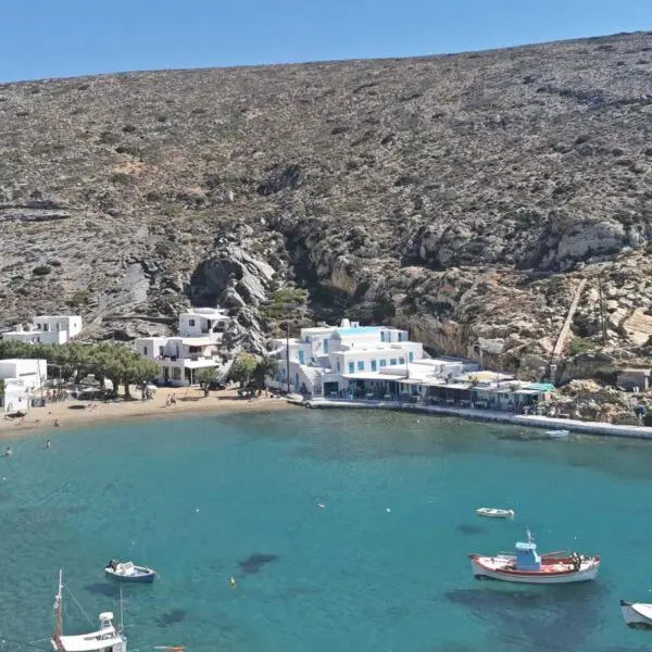 A small coastal village on the island of Sifnos with white buildings is set against rocky hills. Boats are moored on the calm turquoise sea near the sandy shore. A perfect spot for any travel guide, it offers serene views and a peaceful retreat.