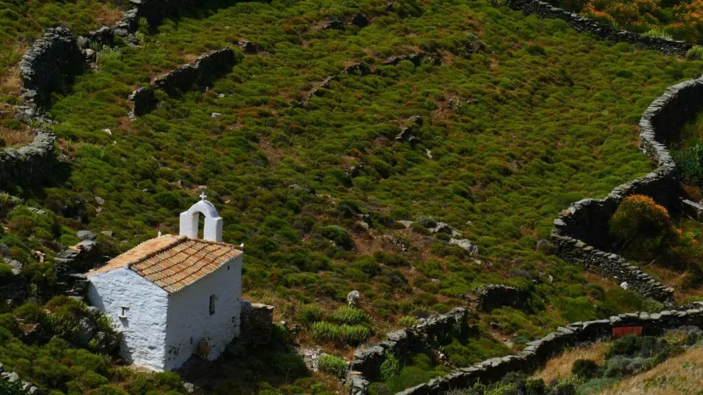 A small white chapel with a tiled roof is nestled among the green terraced hillsides of Kythnos Island, surrounded by stone walls. Perfect for any travel guide seeking hidden gems.