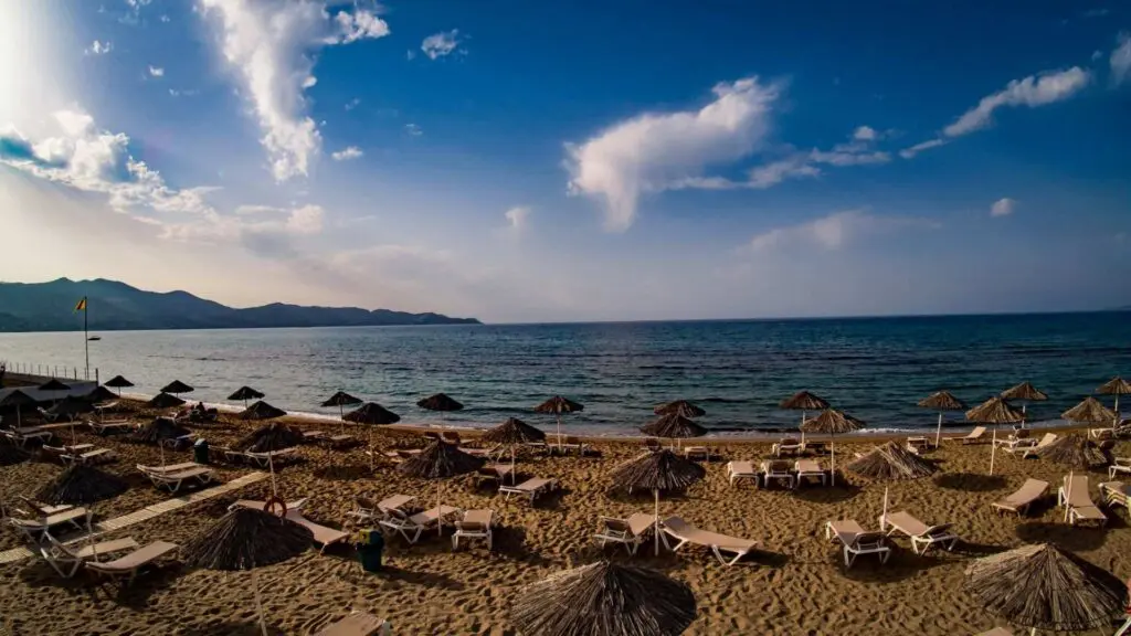 Sandy beach with rows of empty sun loungers and straw umbrellas facing the calm sea under a blue sky with few clouds, reminiscent of Heraklion beaches. Hills are visible in the distance.