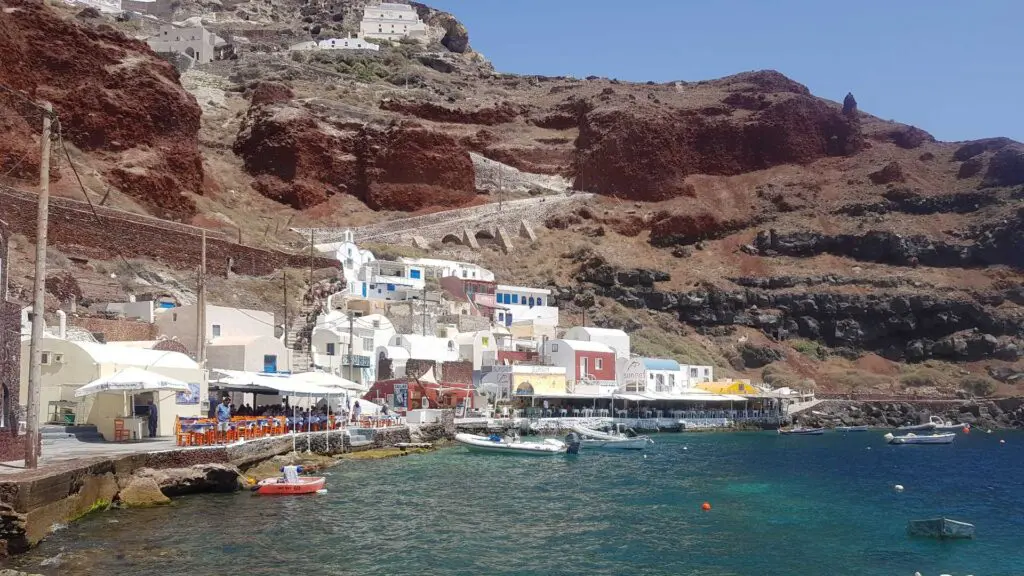Coastal village with white and colorful buildings set against rocky cliffs, boats docked by a clear blue sea, reminiscent of a Santorini ferry port.