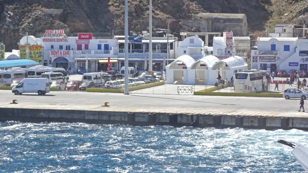 Seaside view of the busy Santorini ferry port, with rental car facilities, parked vehicles, and a few people near the waterfront. Waves can be seen crashing against the shore.