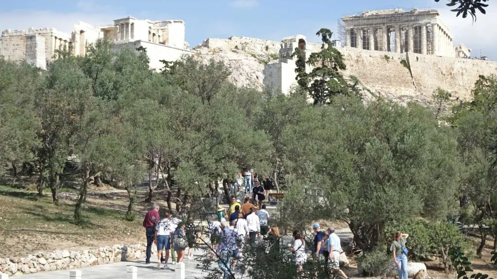 Visitors stroll towards the Acropolis, surrounded by greenery and ancient structures under a clear sky, where historical insights await.