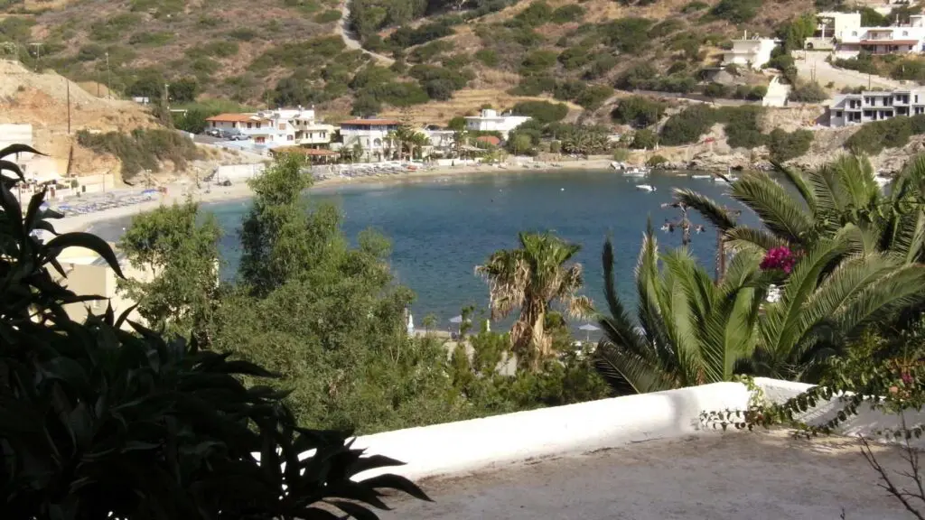 Bay view with clear water, surrounded by lush greenery and hillside houses reminiscent of Heraklion beaches. A few small boats gently glide across the water.