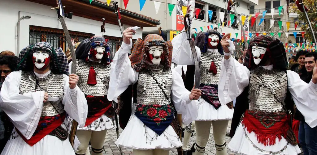 Five people in traditional Greek costumes with white masks and metallic chest armor stand in a festive street setting adorned with colorful flags, embodying the vibrant spirit of Apokries.