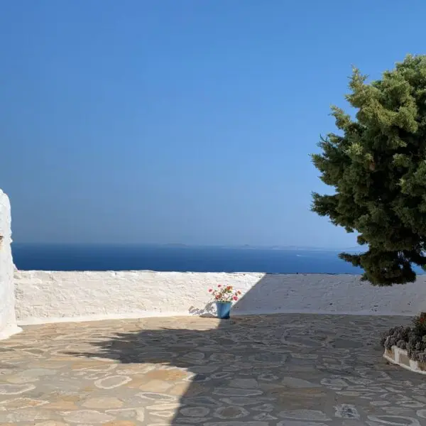 A sunny courtyard with stone paving, whitewashed walls, potted plants, and a large tree overlooks the calm blue sea of Sikinos Island.
