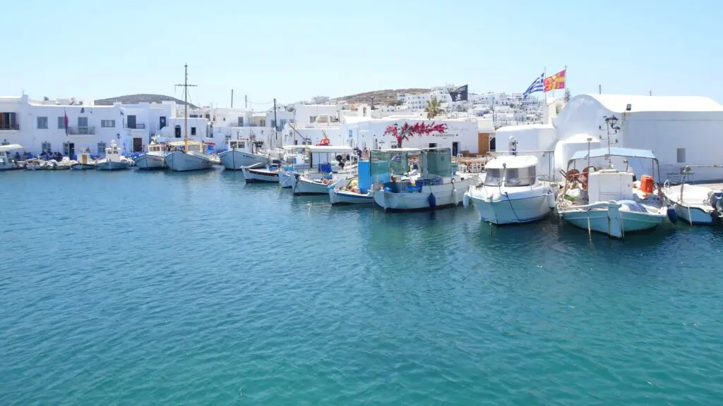 A serene harbor on Paros with white buildings in the background and several small boats moored on calm blue water under a vast, clear sky, just as highlighted in the island guide.
