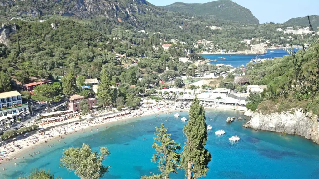 From a traveler's aerial view, this turquoise bay gleams invitingly, its sandy beach fringed with trees and charming buildings. Hills lush with greenery envelop the area, offering a perfect start for your Greek island-hopping itinerary.