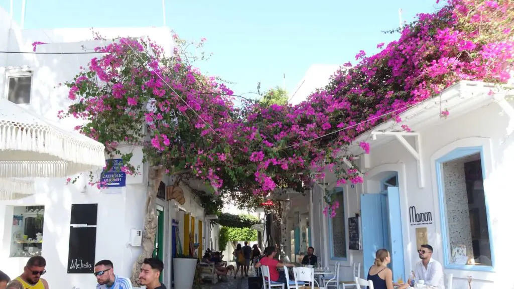 Amidst the charming white-walled town, travelers enjoy leisurely moments at outdoor cafes beneath bougainvillea-clad arches. The bright blue sky completes this idyllic Greek island-hopping scene.