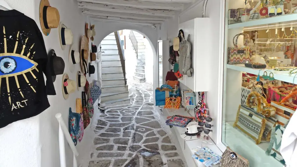 Narrow alleyway with displayed hats, bags, and trinkets under a white arch; a shop brimming with items on the right. White stairs lead upward in the background, reminiscent of an island guide through Paros' charming streets.
