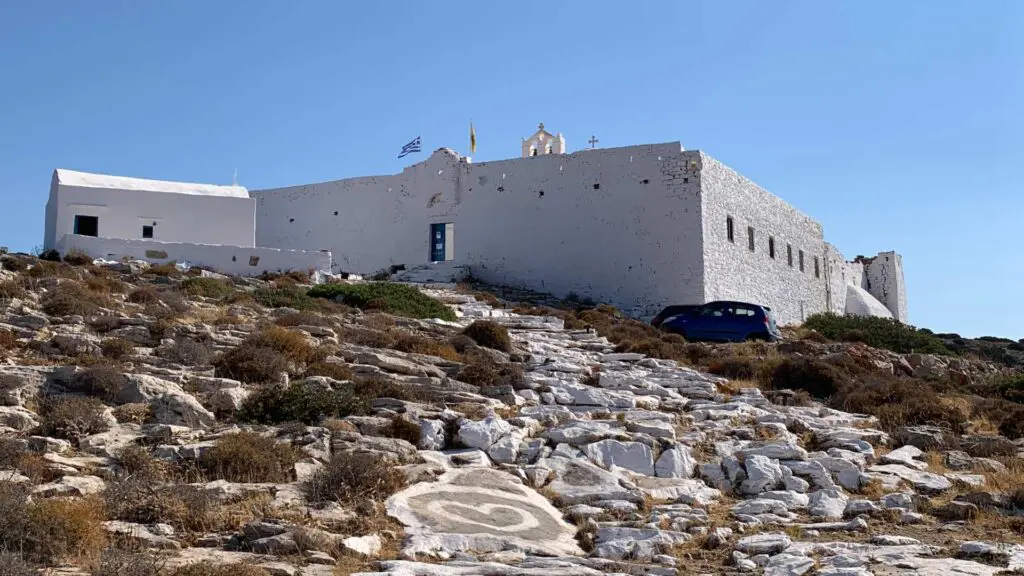 A white stone building perched on Sikinos Island features a sloped pathway and dry vegetation. The Greek flag flutters proudly above.