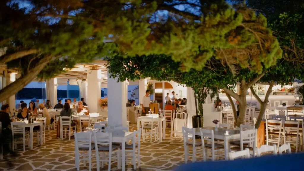 Located in a charming outdoor setting reminiscent of Paros, this restaurant features white tables and chairs nestled under trees, filled with diners enjoying the evening. The candlelit ambiance and natural decor craft a cozy atmosphere, perfect for those choosing the right island dining experience.