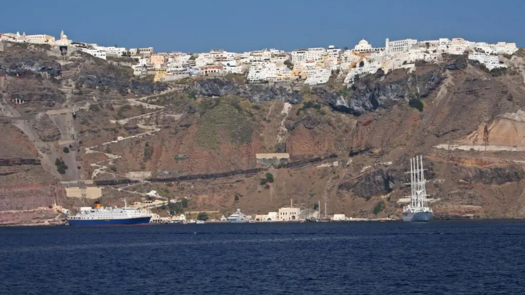 A hillside town with white buildings perches above a rocky cliff, reminiscent of Santorini. Nearby, two ships are docked at the ferry port by the shoreline, embraced by clear blue water.
