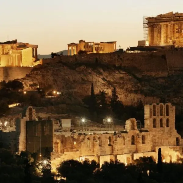 The image offers an insider’s guide to the Acropolis of Athens at dusk, with illuminated ancient structures like the Parthenon and the Odeon of Herodes Atticus in the foreground, providing historical insights into this breathtaking masterpiece.