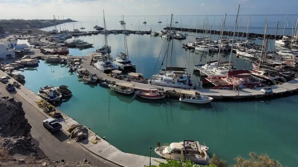 The circular marina, reminiscent of the Santorini ferry port, is a serene scene with various sailboats and yachts docked, surrounded by calm water and a rugged rocky coastline.