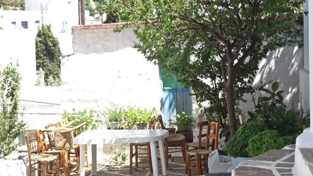 In the charming village of Lefkes, Paros, Greece, an outdoor patio features a white table surrounded by wooden chairs under a tree. Sunlight dances through the leaves, casting gentle shadows on the ground.