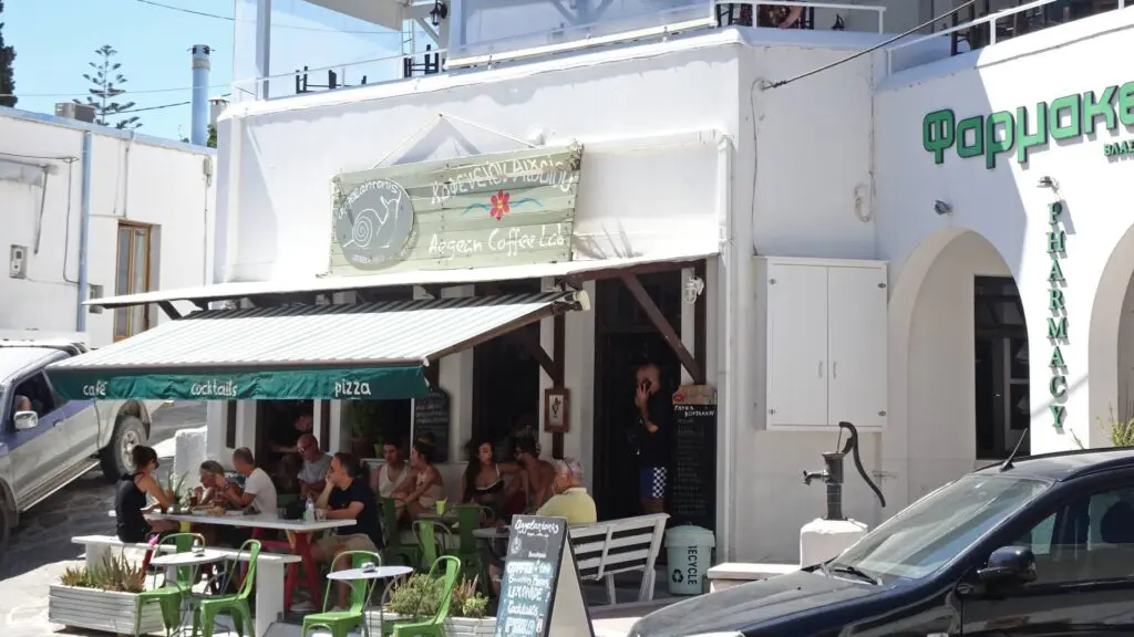 People sitting outside a cafe in Lefkes, Paros on green chairs, under a hanging sign against white walls. A green awning offers shade. A car is parked in front, and a pharmacy is visible to the right.