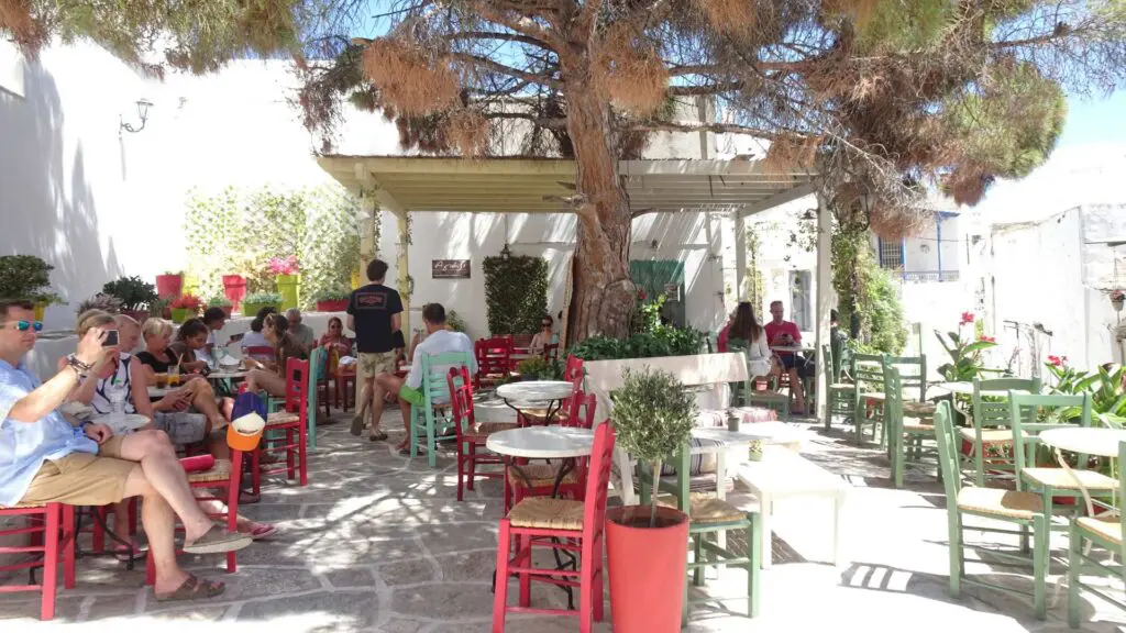 Outdoor café in Lefkes, Paros, Greece, with people sitting at colorful tables under a large tree. Some are chatting, while others use phones.
