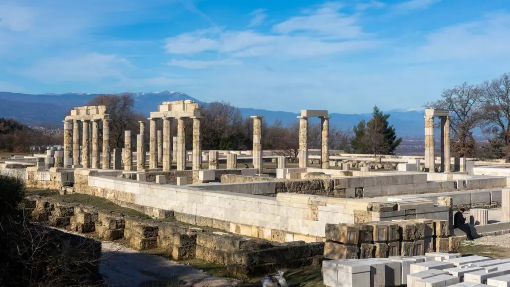 Ancient stone ruins with columns and bases, nestled in the picturesque landscape of Vergina, are set against a backdrop of trees and distant hills under a blue sky.
