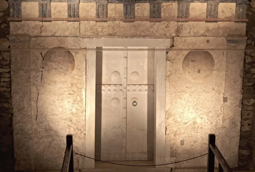 The ancient stone door, reminiscent of Vergina's archaeological wonders, features circular carvings and is set in a weathered wall adorned with decorative elements above. A rope barrier stands in front to protect its historical significance.