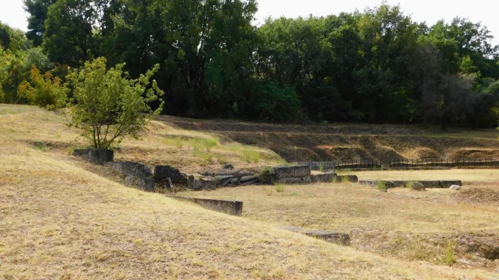 Ancient stone ruins of Vergina are nestled amid lush grass and trees, set on a sloped landscape under a clear sky.