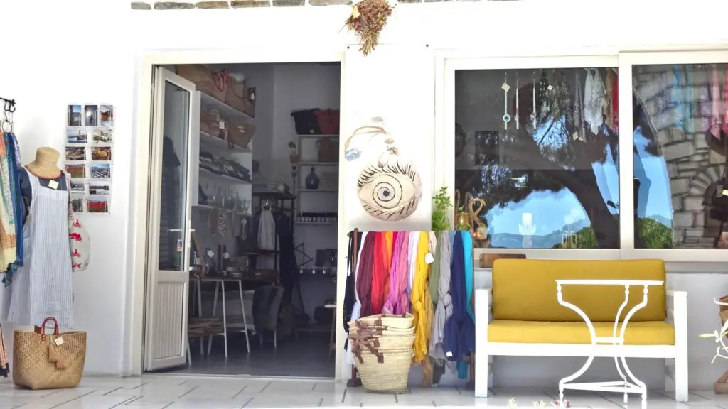 Outdoor view of a charming shop in Lefkes, Paros, Greece, with a white exterior. It features colorful scarves, a yellow bench, and wicker baskets. The shop's door and window reveal an inviting interior adorned with hanging clothes and accessories.