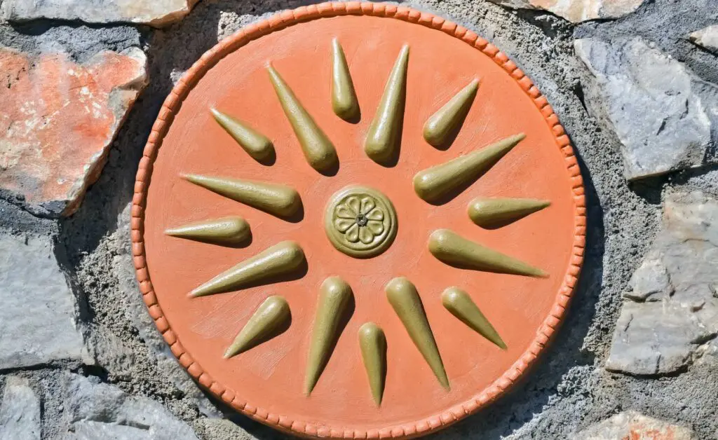 Circular terracotta plaque with radial, pointed designs and a small central floral motif reminiscent of the Vergina Sun, mounted on a stone wall.