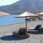 Beach scene with straw umbrellas and sun loungers on the rocky shore of Pomos, Cyprus, overlooking the blue sea and mountainous background. Several people are seated under umbrellas, enjoying the serene view.