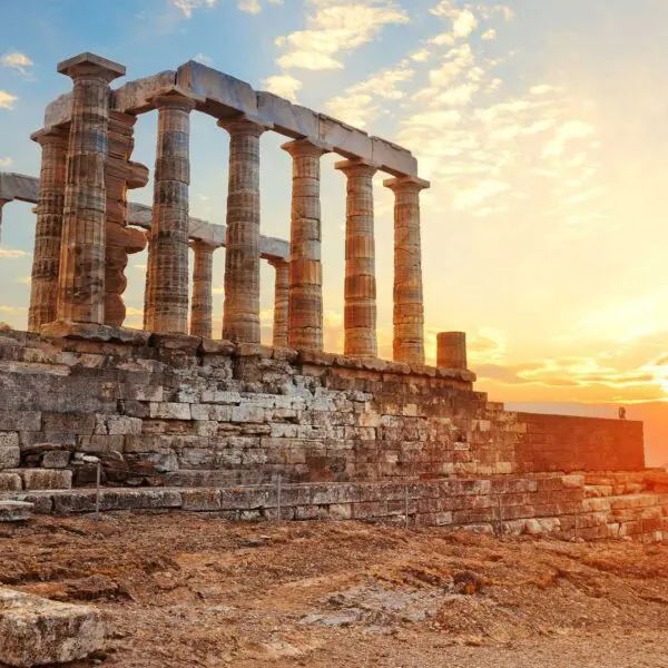 The ancient stone columns of the Temple of Poseidon at Cape Sounion, Greece, stand against a sunset sky with the sea and a distant island in the background—one of the must-see places to visit in Greece.